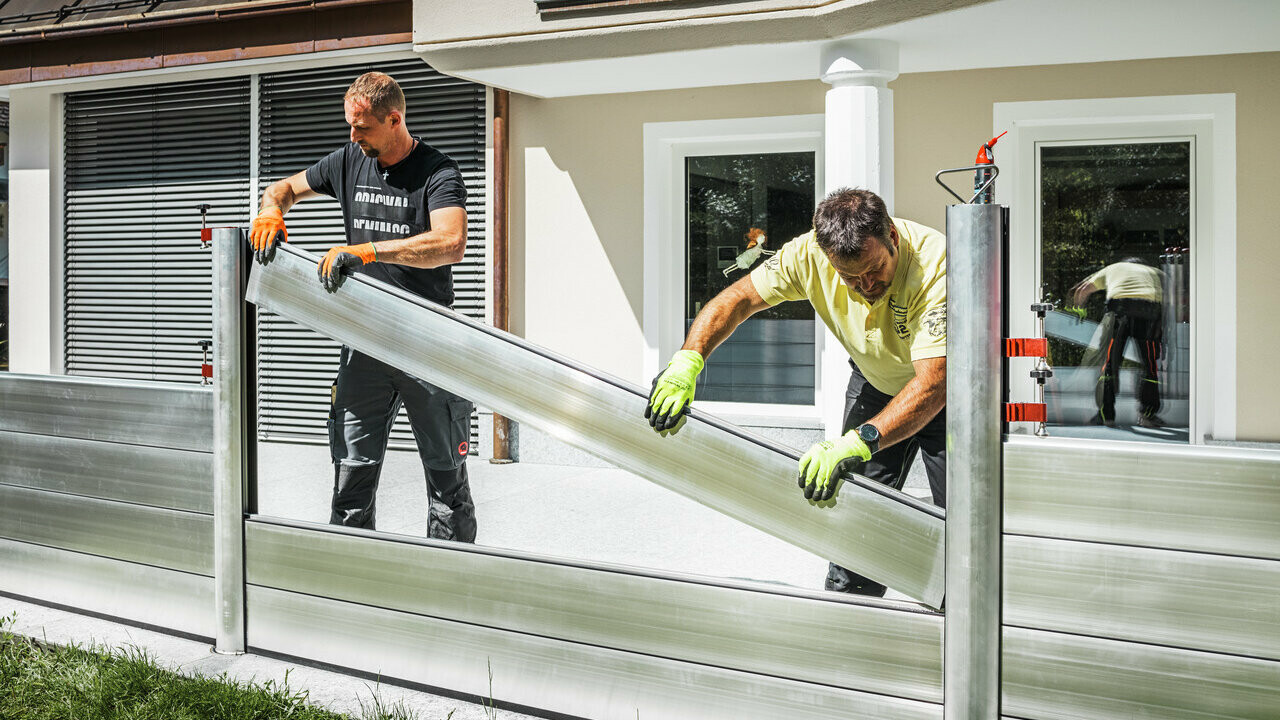 Two men can be seen setting up the PREFA flood protection system and inserting a dam beam into the profile rails.