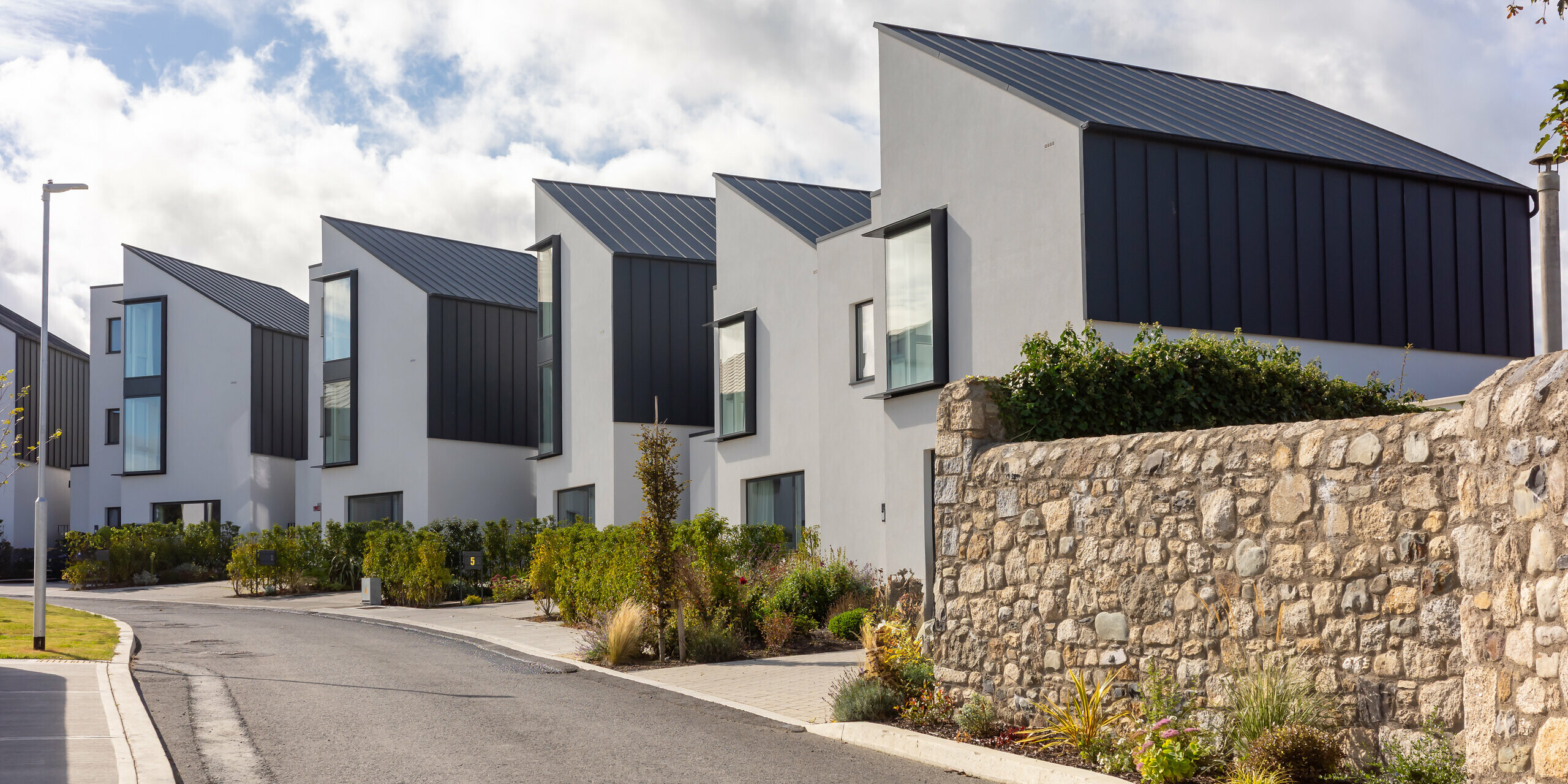 Watson Place terraced housing development with striking sloping roofs and continuous PREFALZ standing seam cladding in P.10 anthracite. The dark aluminium surfaces create a striking contrast to the light-coloured façades and natural stone walls. The durable PREFA aluminium provides reliable weather protection and gives the modern architecture a clear, high-quality appearance.