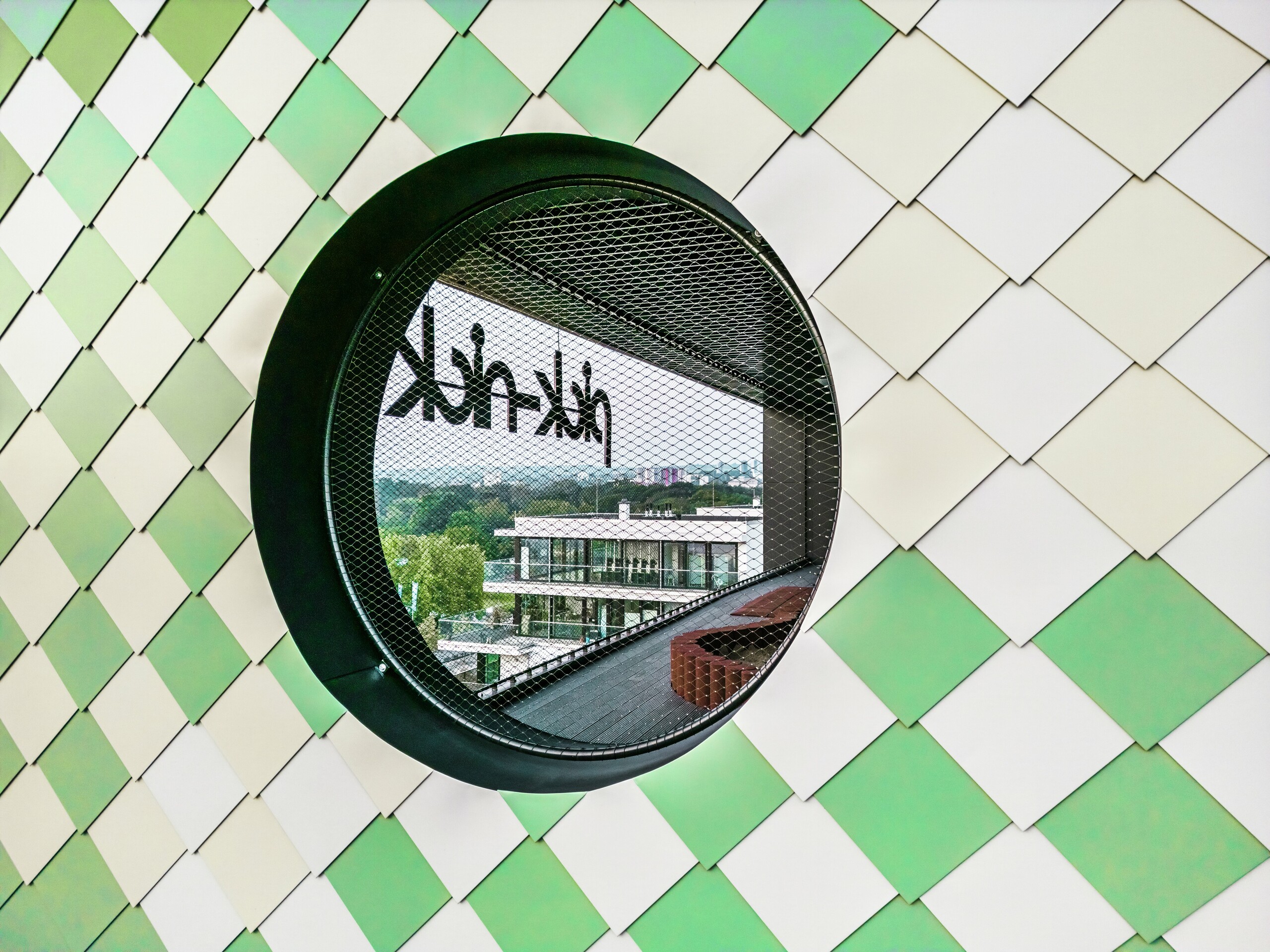 Detailed view of the PREFA aluminium façade on the ‘Pick-Nick’ residential and office building in Dresden. The focal point is a round window with a black frame and integrated grating. When looking through the round window, the mirror-inverted word ‘Pick-Nick’ can be seen. The façade combines design, functionality and lasting protection in perfect harmony.