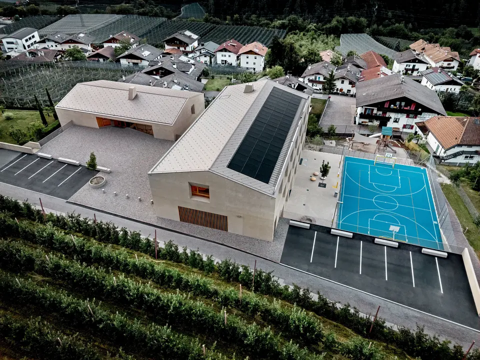 Zračna snimka obrazovnog centra Riffian u Južnom Tirolu s dječjim vrtićem, osnovnom školom i sportskim igralištem. PREFA krovni rombovi dimenzija 44 × 44 u boji P.10 bronca tvore preciznu i dugotrajnu aluminijsku krovnu oblogu koja se skladno uklapa u alpski krajolik. Moderna arhitektura ističe se otpornošću na vremenske uvjete i čistim dizajnom do najsitnijih detalja.