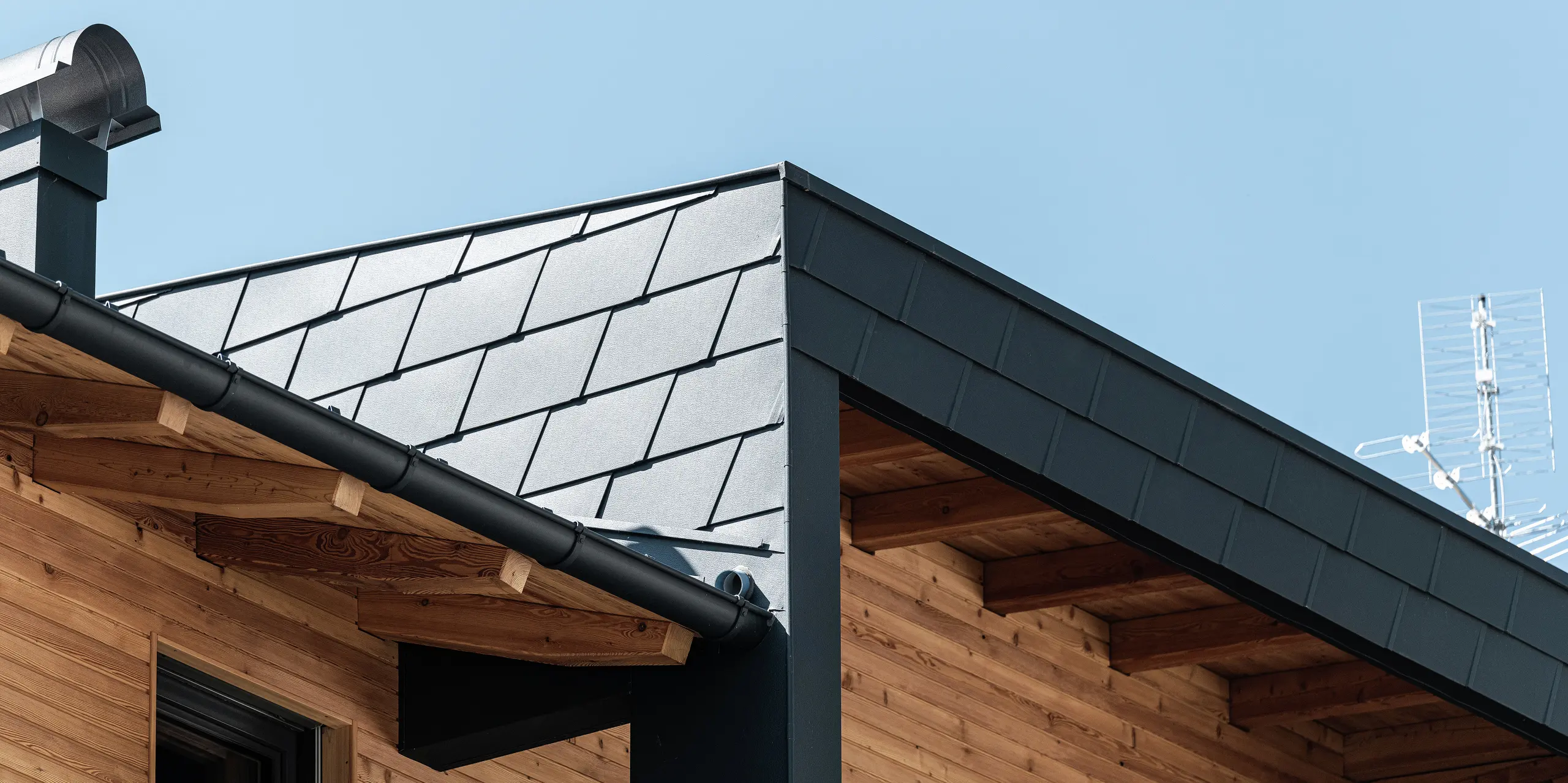 Detailansicht der Dachecke mit PREFA Dachschindeln DS.19 in der Farbe P.10 Anthrazit und der Fassadenverkleidung des überdachten Balkons. Die Kombination von Aluminium und Holz setzt ästhetische Akzente.