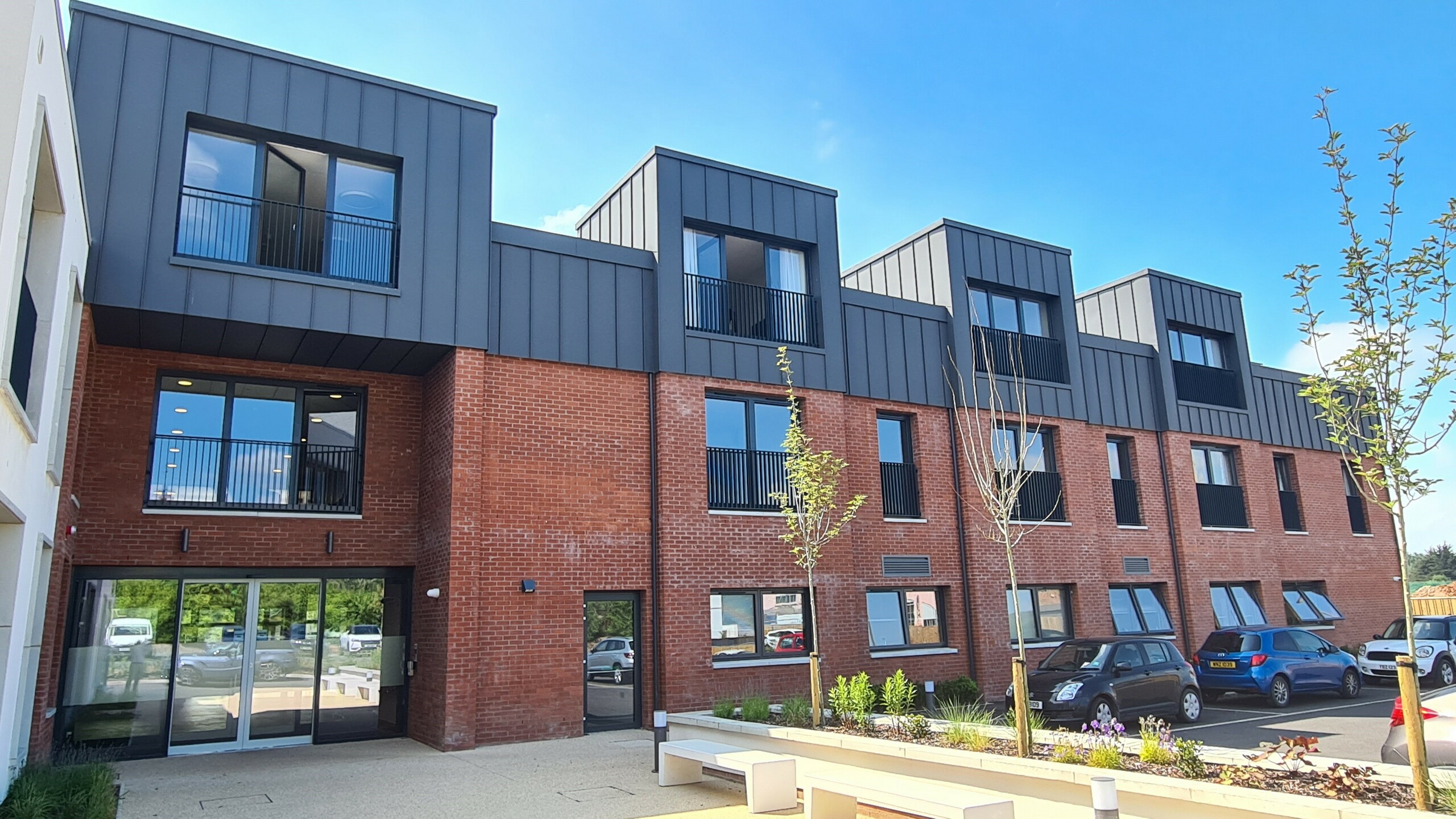 Front view of Harberton Hall in Belfast, Northern Ireland. The standing seam roof made of PREFALZ P.10 dark grey perfectly complements the brick façade. Large windows and flat roof dormers emphasise the innovative design of the building for assisted living and create a welcoming atmosphere. The glass entrance door provides insights into the internal facilities.