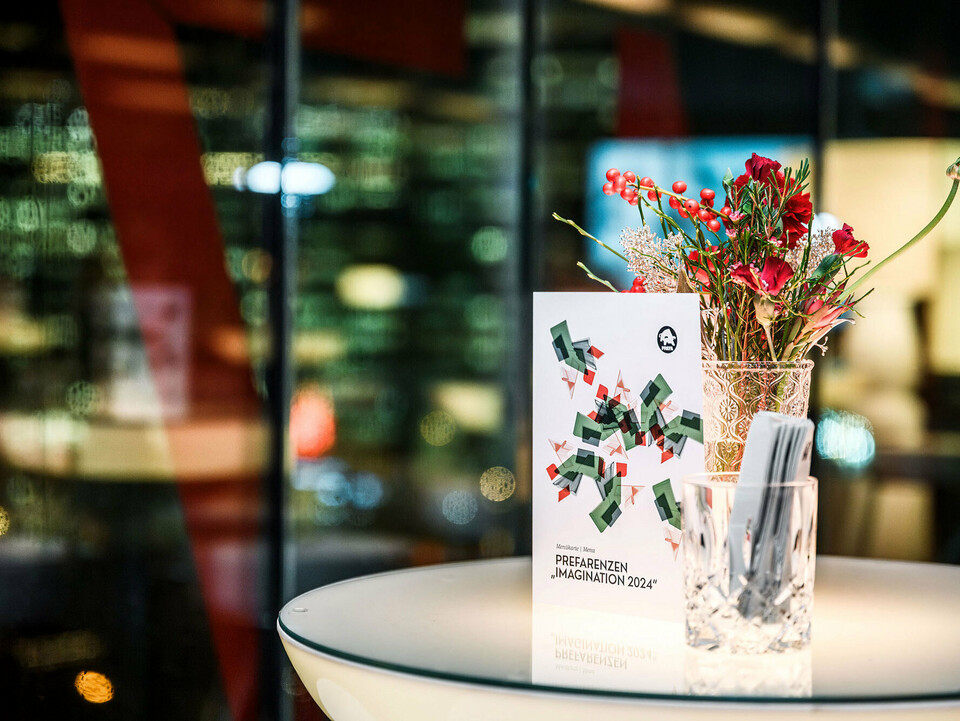 Normal view of a PREFARENZEN event 2024 invitation on a table before a flower arrangement in a glass jug, with a blurred background behind it.