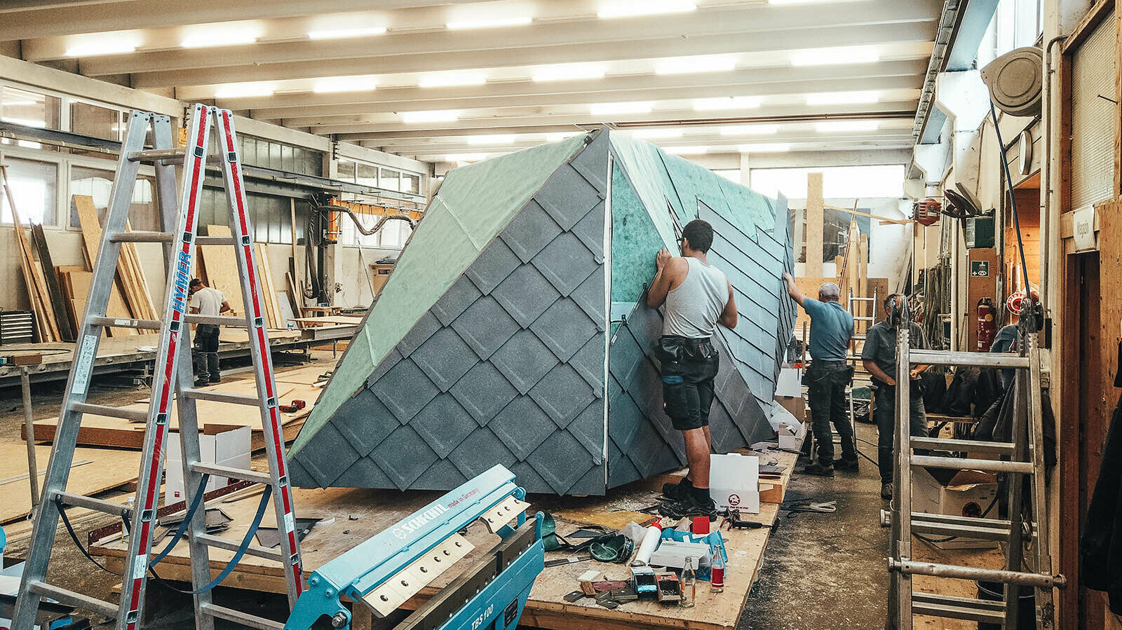 The picture shows the plumbers at work. PREFA rhomboid roof tiles 29x29 are being attached to the building.