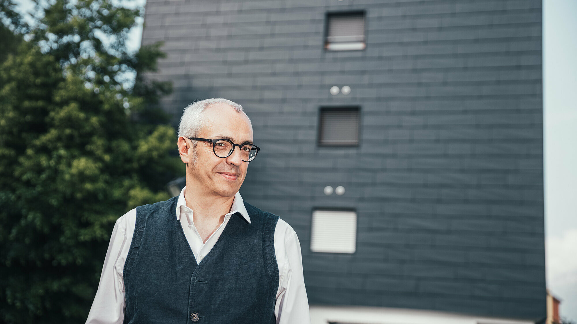 Portrait of architect Manuel Benedikter in front of the thermally renovated residential building with the anthracite-coloured PREFA façade.