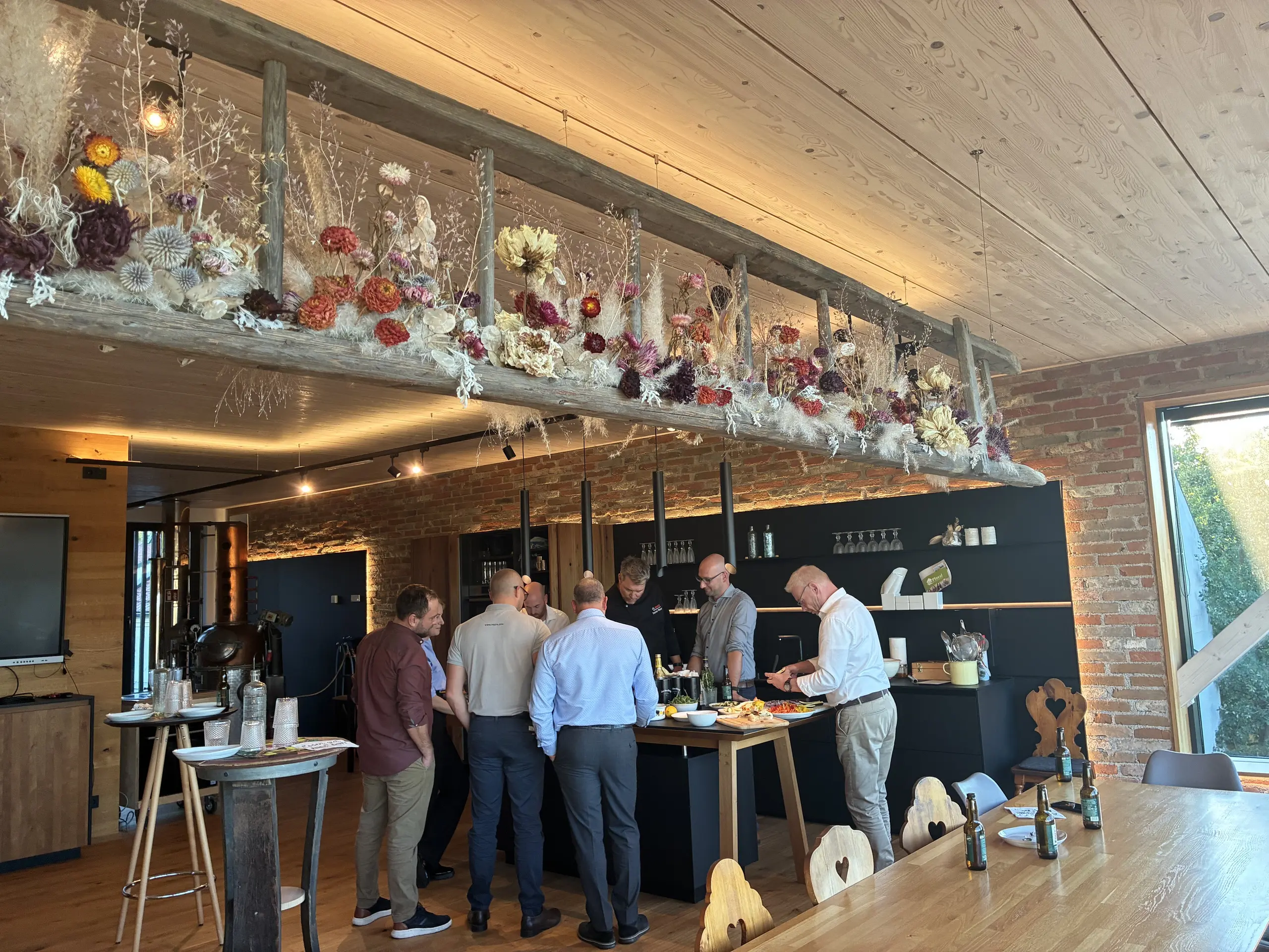 A group of people are standing around a kitchen island during a get-together in a cosy room with wooden and brick walls and floral decorations.