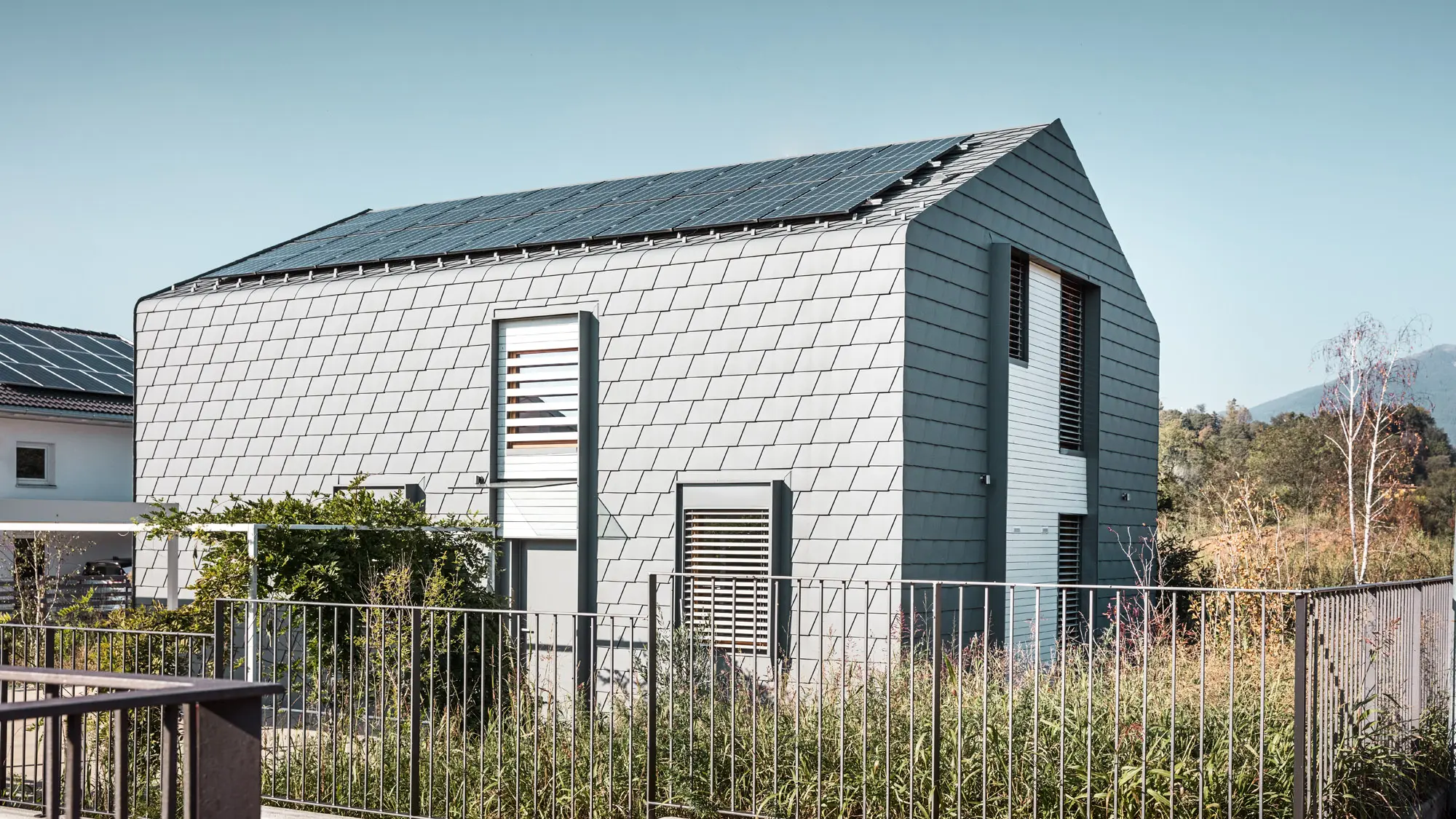 Lateral view of "Casa Longaretti" with a green area and fence, the round transition between roof and façade cis clearly visible, just like the light grey shingle façade.