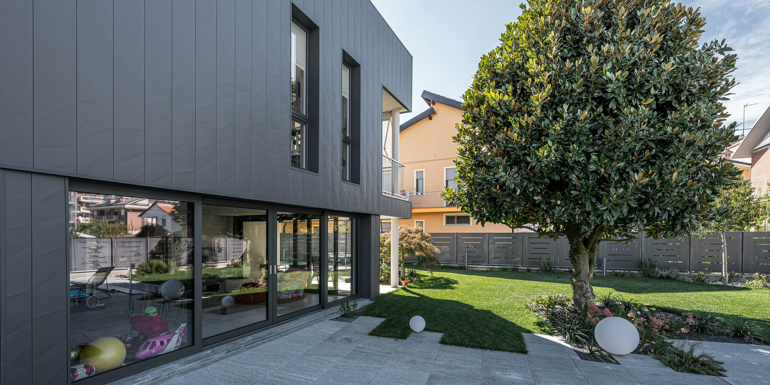 Detaillierte Seitenansicht eines modernen Einfamilienhauses in Savigliano, Italien, hervorgehoben durch eine stilvolle PREFA Siding.X Aluminiumfassade in P.10 Dunkelgrau. Die großzügigen Fenster und Glasfronten maximieren die natürliche Beleuchtung und verbinden das Innere nahtlos mit dem gepflegten Außenbereich. Diese Blechfassade repräsentiert eine Kombination aus Nachhaltigkeit und zeitgemäßem Design, wodurch das Gebäude sowohl ästhetisch ansprechend als auch funktional ist.