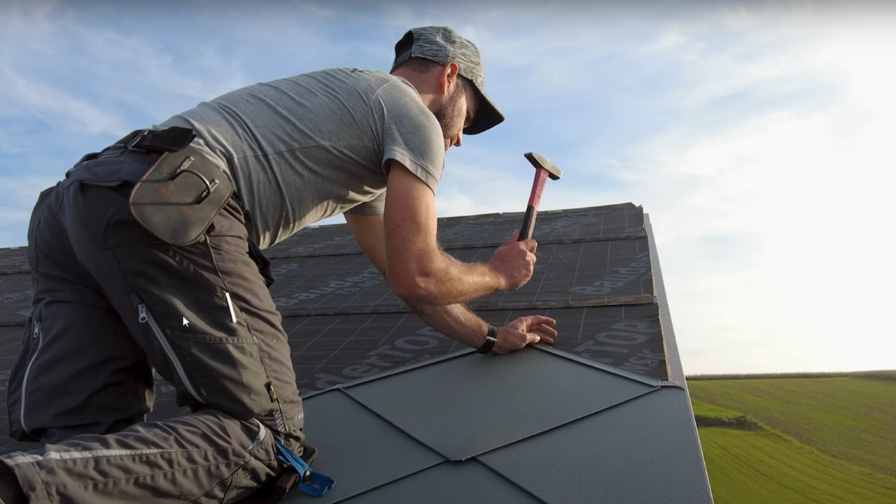 Installer in action: The owner of the company ‘Der Blechmann’ uses a hammer to precisely install PREFA rhomboid roof tiles 44 × 44. The precision of the installation can be seen in the rhomboid roof tiles already installed at the bottom of the image. The man is wearing a cap and kneeling on the roof with his back to the camera. The clear sky and green landscape can be seen in the background. The harmonious background emphasises the symbolism of the image: sustainability and durability in craftsmanship with high-quality PREFA aluminium products.