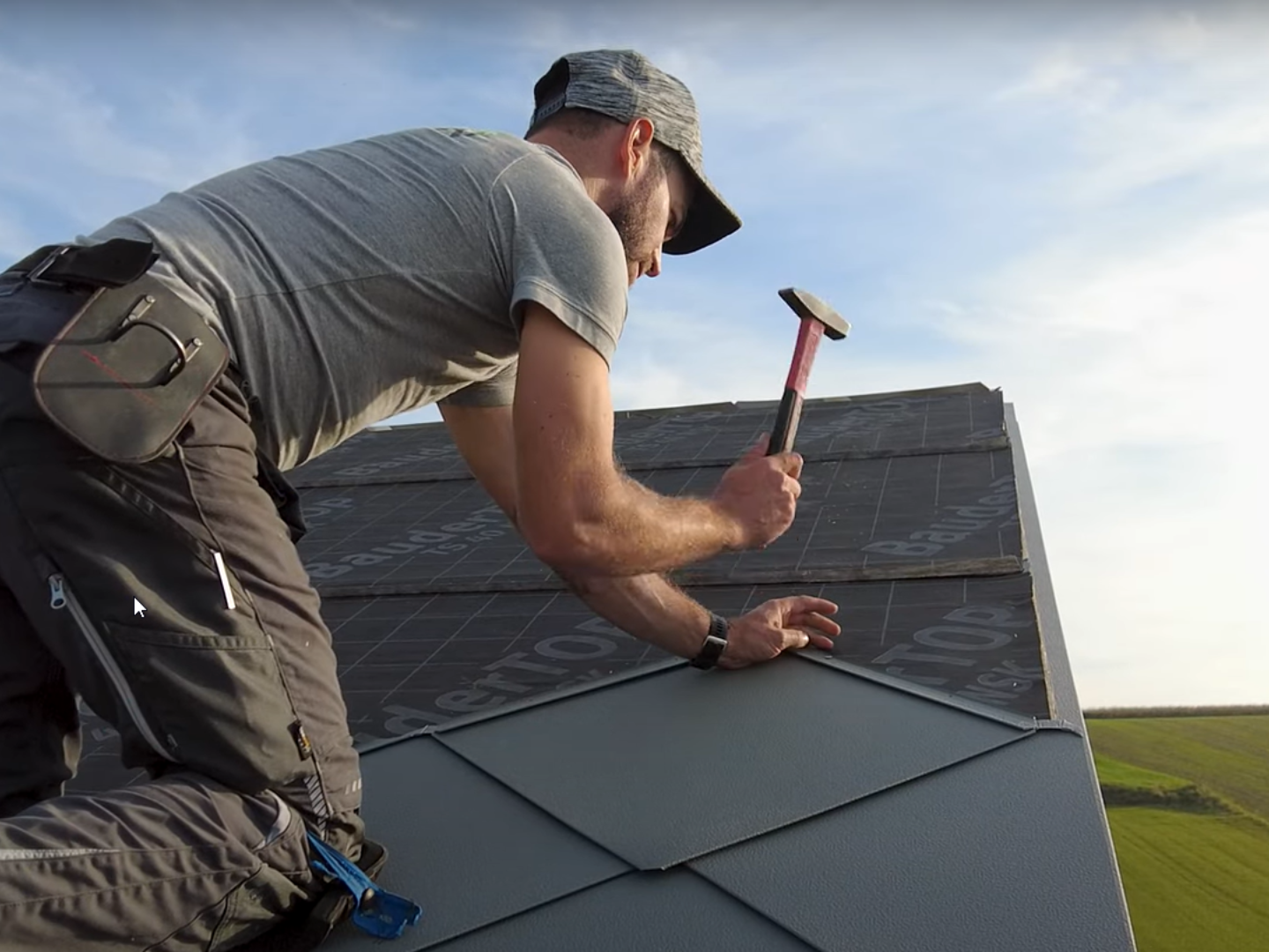 Installer in action: The owner of the company ‘Der Blechmann’ uses a hammer to precisely install PREFA rhomboid roof tiles 44 × 44. The precision of the installation can be seen in the rhomboid roof tiles already installed at the bottom of the image. The man is wearing a cap and kneeling on the roof with his back to the camera. The clear sky and green landscape can be seen in the background. The harmonious background emphasises the symbolism of the image: sustainability and durability in craftsmanship with high-quality PREFA aluminium products.