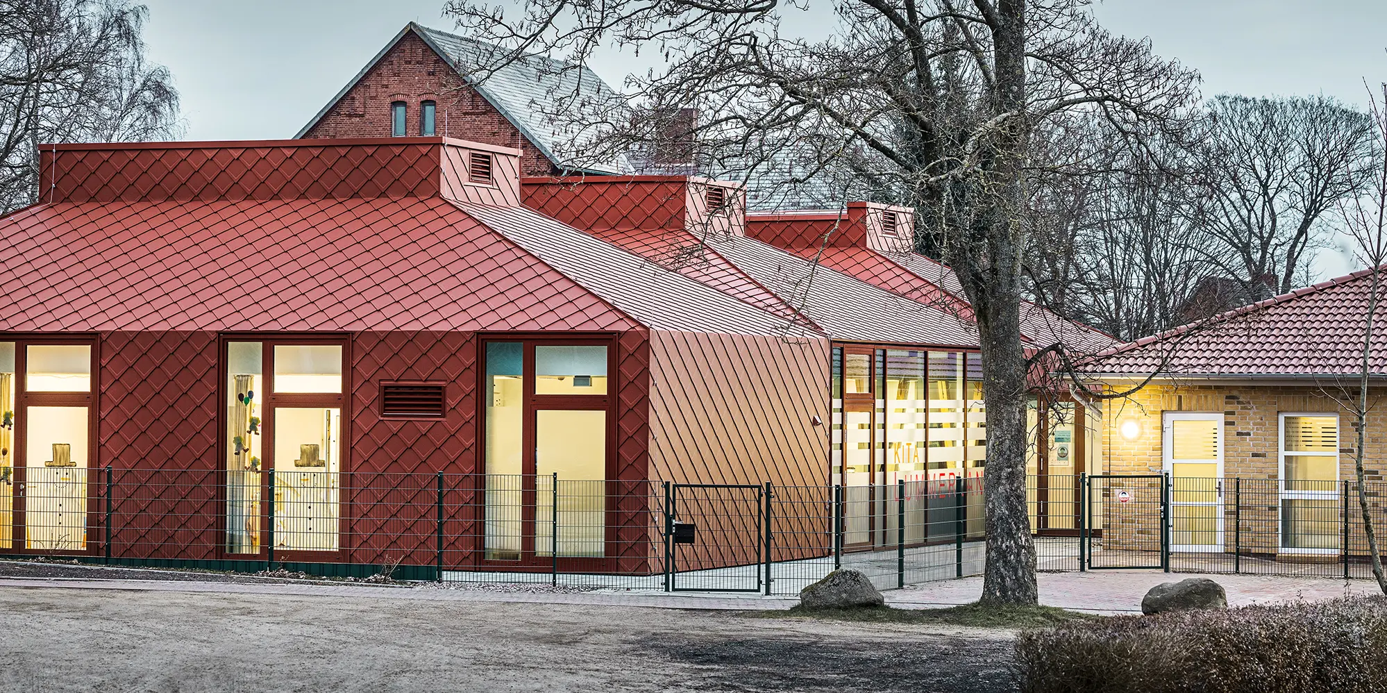 Modernes Kindergartengebäude mit auffälliger Fassade und Dacheindeckung aus rautenförmigen Aluminiumplatten in Oxydrot. Die Architektur kombiniert traditionelle und moderne Elemente mit großen Fenstern und klaren Linien. Das Gebäude ist von Bäumen umgeben, im Vordergrund verläuft ein Zaun, und die Beleuchtung im Inneren schafft eine warme Atmosphäre in der Dämmerung.