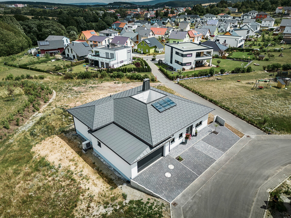 Moderne Architektur trifft Funktionalität: Ein Bungalow in Ilmenau, Thüringen, mit einem beeindruckenden PREFA Aluminiumdach bestehend aus der Dachplatte R.16 in P.10 Dunkelgrau. Die Dachlandschaft zeigt ein zentrales Atrium und einen markanten Schornstein mit dem charakteristischen PREFA Stier, ergänzt durch ein langlebiges PREFA Dachentwässerungssystem mit Ablaufrohr und Hängerinne. Die Garage fügt sich nahtlos in das Gesamtbild ein, verkleidet mit PREFALZ in passendem Dunkelgrau.