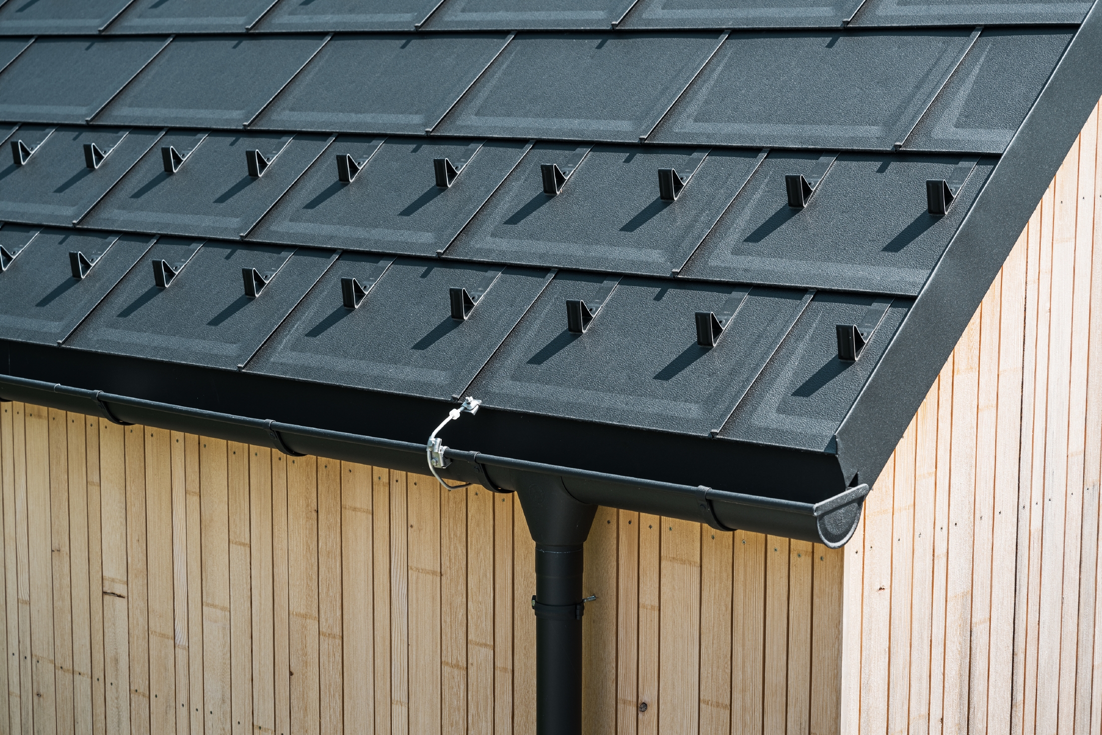 Close-up of PREFA roof tile R.16 with snow guards, gutter and downpipe in black P.10 incl. lightning protection and a vertical wooden façade