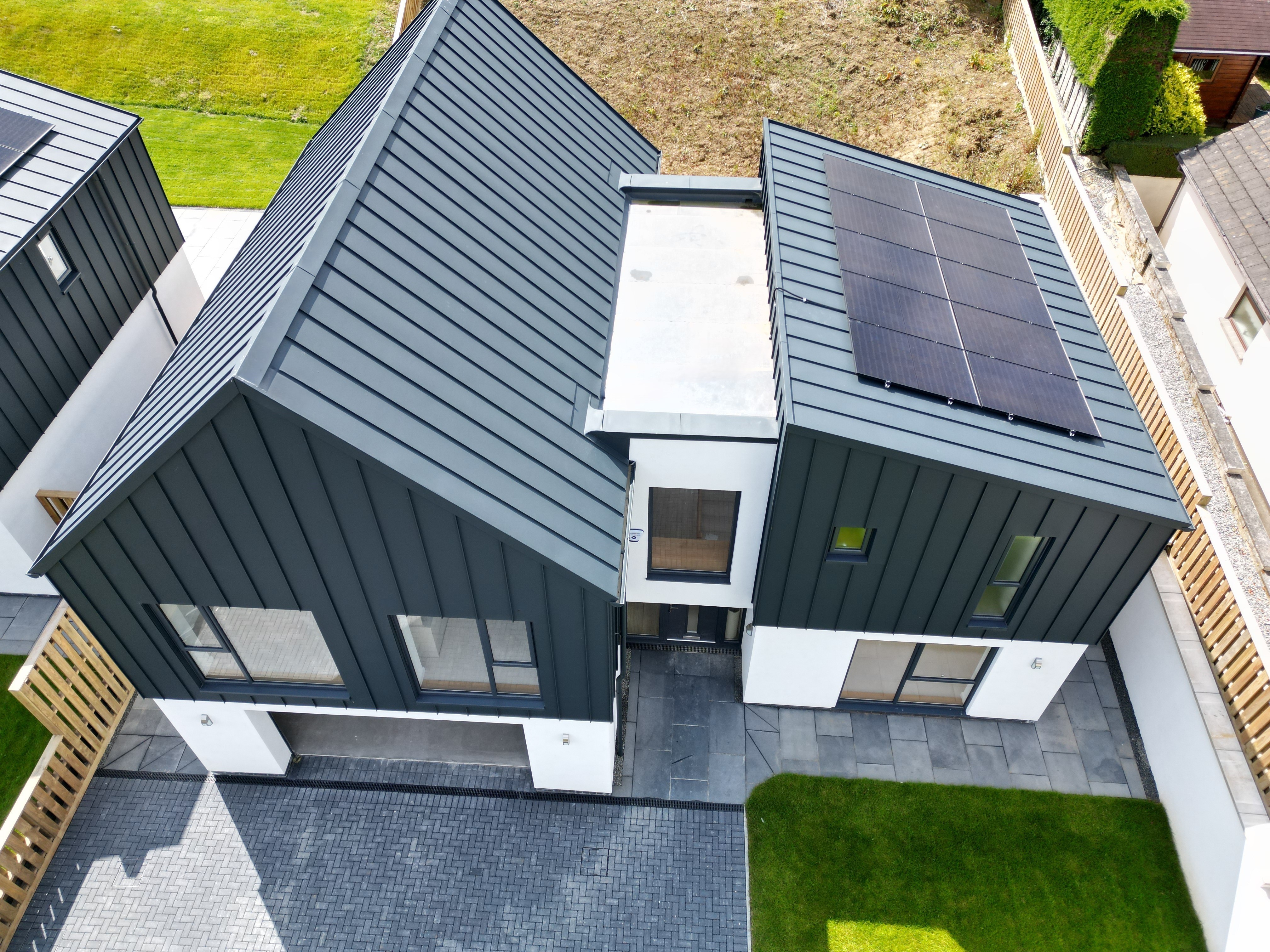 A bird's eye view of a detached house in Bridgerule, Devon, with PREFALZ P.10 anthracite as a standing seam covering on the roof and façade. The modern architecture combines durable aluminium with aesthetic longevity, complemented by a solar energy system. A gable roof towers over the garage entrance, while the right-hand part of the building has a pent roof. Between the roofs, a building bridge connects the two structures.